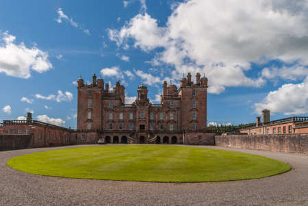 Holm of Drumlanrig, Scotland, UK - June 18, 2012: Front facade with double staircase and clock tower of free standing pink sand stone Drumlanrig Castle on its higher plateau. Circular green lawn up front.のeditorial素材