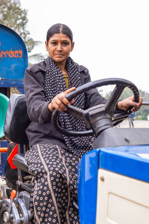 Angadihalli, Karnataka, India - November 2, 2013: Closeup of young Hindu woman in marine blue dress drives a blue tractor pulling corn threshing machine on street.のeditorial素材