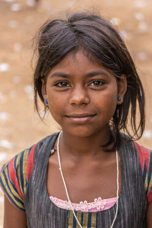 Angadihalli, Karnataka, India - November 2, 2013: Closeup of young girl with black hair and blue pins in her ears smiles and wears colorful shirt and dress. Beige background.のeditorial素材