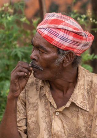 Angadihalli, Karnataka, India - November 2, 2013: Closeup of face of man with red and white turban and brown shirt picking his teeth. Looks away. Green background.のeditorial素材