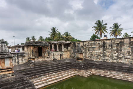 Belur, Karnataka, India - November 2, 2013: Chennakeshava Temple. Brown stone dilapidated shrine in corner of the grounds adjacent to green water tank. Darkened steps with elephant statues and green of palm trees above.のeditorial素材