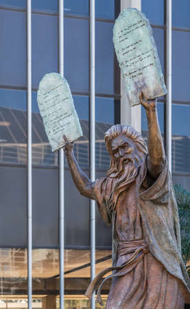 Garden Grove, California, USA - December 13, 2018: Crystal Christ Cathedral. Closeup of Bronze statue of Moses putting the ten commandments of two tables in the air. Some green foliage, Tower of hope in back.のeditorial素材