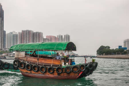 Hong Kong, China - May 12, 2010: Green roofed brown wooden sampan on greenish waters of central Harbor. High rise buildings as background under silver sky.のeditorial素材