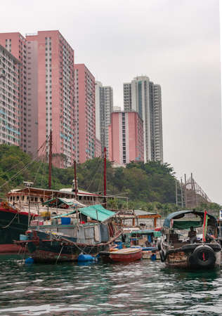 Hong Kong, China  - May 12, 2010: Brown wooden ferry sampan and group of houseboats and sloops docked in  harbor with white tall apartment buildings on shore. Silver sky.のeditorial素材