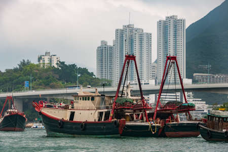 Hong Kong, China  - May 12, 2010: Black and red fishing vessels docked in  harbor with white tall apartment buildings on shore. Silver sky over green hill. Bridge span over boats.のeditorial素材
