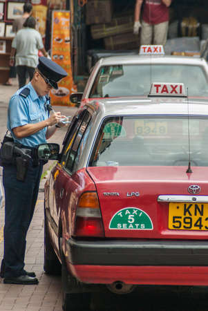 Hong Kong Island, China  - May 12, 2010: A traffic cop in blue writes a red taxi cab up for an infraction near Stanley Market. Faded shops and vendors in back.のeditorial素材