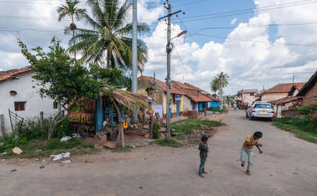 Belavadi, Karnataka, India - November 2, 2013: Two little boys play in the street in front of Veera Narayana Temple. Red roofed houses, cars, under blue sky with heavy cloudscape. Shop owner in shack looking.のeditorial素材