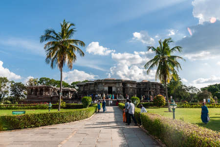 Halebidu, Karnataka, India - November 2, 2013: Brown stone flat Hoysaleswara Temple of Shiva in its green garden with palm trees and plenty of devotees in colorful dress under blue sky with white clouds.のeditorial素材