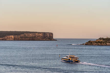 Sydney, Australia - February 12, 2019: North and South Head cliffs at gate between Tasman Sea and Sydney Bay during sunset. Cloudless pale sky. Gray water. Warm brown rocks and a yellow ferry boat.のeditorial素材