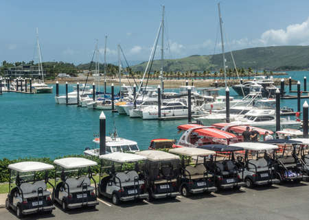 Hamilton Island, Australia - February 16, 2019: Row of golf carts as regular street vehicles parked at yacht harbor on marina. Azure water, white boats, green hills, blue sky.のeditorial素材
