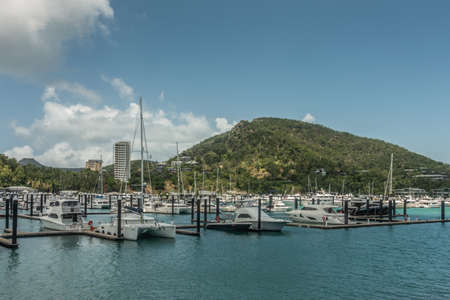 Hamilton Island, Australia - February 16, 2019: Wider shot of the marina with its yacht harbor full of white boats. Green hill in the back under blue sky with some white clouds.のeditorial素材