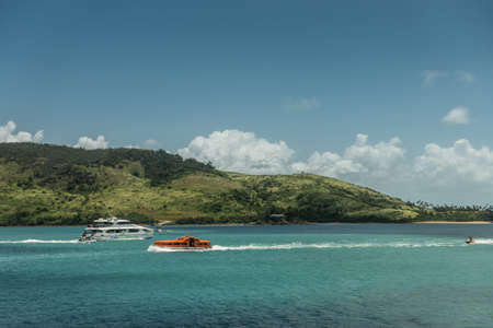 Hamilton Island, Australia - February 16, 2019: Jetski and two boats just outside the marina. Green hill in the back under blue sky with some white clouds.のeditorial素材