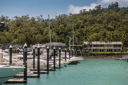 Hamilton Island, Australia - February 16, 2019: Bakery, captains and other eateries on Front Street behind row of white boats moored in marina. Back is greeen foliage of forest under blue sky. Azure water.のeditorial素材