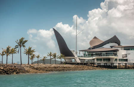 Hamilton Island, Australia - February 16, 2019: Black and white Iconic Yacht Club House behind azure water and under blue sky with white cloudscape.のeditorial素材