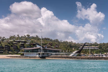 Hamilton Island, Australia - February 16, 2019: Wider shot of Black and white Iconic Yacht Club House, shot from sea, under blue sky and white cloudscape. Green hill with housing in back.のeditorial素材