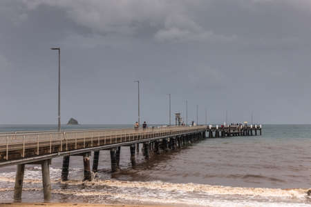 Cairns, Australia - February 17, 2019: Palm cove pier extends into gray Coral Sea under a post rain storm dark sky. Some people on the jetty. Rock Island on horizon.のeditorial素材
