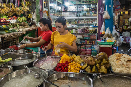 Sihanoukville, Cambodia - March 15, 2019: Phsar Leu Market. Two young women prepare and serve fast food, fruits, pickles and more out of basins and pot. Other stores in back.のeditorial素材