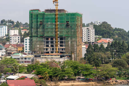Sihanoukville, Cambodia - March 15, 2019: Large highrise construction site wrapped in green with yellow crane in Sihanouk Autonomous Port neighborhood. Green park on side. Shore up front.のeditorial素材