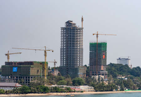 Sihanoukville, Cambodia - March 15, 2019: Shoreline turned into massive construction site of tall high rise buildings under light blue sky, yellow cranes and sandy beach with green trees.のeditorial素材