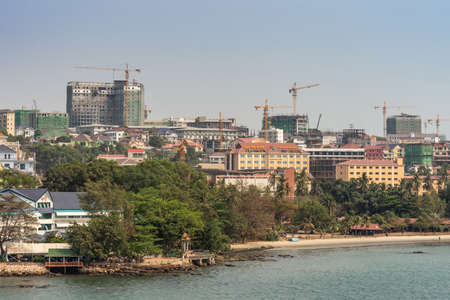 Sihanoukville, Cambodia - March 15, 2019: Town turned into massive construction site of tall high rise buildings under light blue sky, yellow cranes. Sandy beach with green trees and blue water up front.のeditorial素材