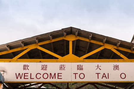 Hong Kong, China - March 7, 2019: Tai O Fishing village. Dual lingual red-on-white yellow sign welcoming visitors to the village, under yellow-beamed roof near creek.のeditorial素材