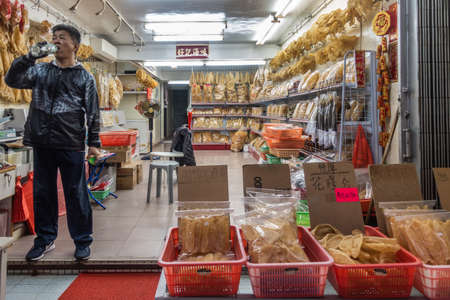 Hong Kong, China - March 7, 2019: Tai O Fishing village. Shop owner is black clothes drinks water while guarding his large dried seafood store, displying all yellowish merchandise on shelfs and in red baskets.のeditorial素材