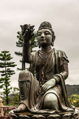 Hong Kong, China - March 7, 2019: Lantau Island. Front Closeup, One of the Six Devas offers flower to Tian Tan Buddha. Bronze statue seen from front with green foliage and rainy sky in back.の写真素材