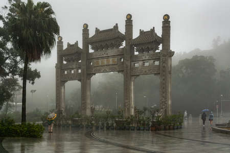 Hong Kong, China - March 7, 2019: Lantau Island. Po Lin Buddhist Monastery. Gray stone ornamental gate under heavy rain blocks the view of the giant Buddha. Some dark green foliage and dense fog.のeditorial素材