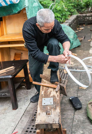 Hong Kong, China - March 8, 2019: Tai Po Market in New Territory. Older cooper or barrel maker fixes metal band into circle at his open air workshop.のeditorial素材