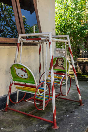 Dusun Ambengan, Bali, Indonesia - February 25, 2019: Family compound. Small children swing for two show images of Hello Kitty and bear. Red an white frame. Wall and green vegetation.のeditorial素材