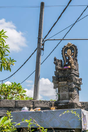 Juga, Bali, Indonesia - February 26, 2019: Sari Pertiwi Wood Carving Business. Gray stone with gold trim Hindu shrine at entrance with small offerings in front under blue sky. Throne like composition.のeditorial素材