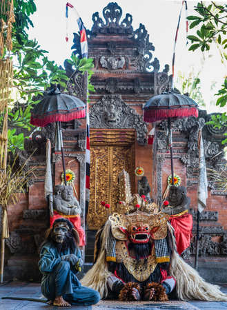 Banjar Gelulung, Bali, Indonesia - February 26, 2019: Mas Village. Play on stage setting. Monkey figure in blue jumper sits besides rised dragon in front of split gate of temple. Reds brake browns.のeditorial素材