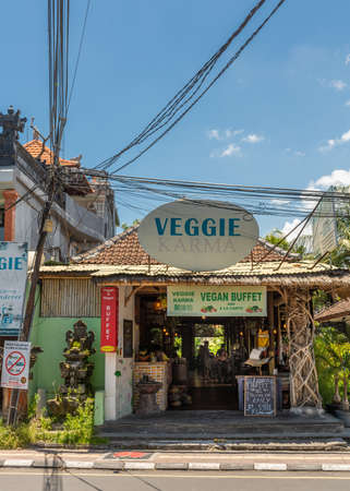 Ubud, Bali, Indonesia - February 26, 2019: Downtown Pedang Tegal, Ji. Hanoman street, commercial district. Front of Veggie Karma, vegan restaurant. Hindu statues, electrical cables in blue sky, green vegetation around.のeditorial素材