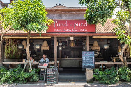 Ubud, Bali, Indonesia - February 26, 2019: Downtown Pedang Tegal, Ji. Hanoman street, commercial district. Closeup of front of Pundi-Pundi coffee and juice bar with guests looking to street. Green foliage, some reds and lots of beige and brown.のeditorial素材