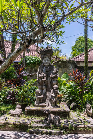 Ubud, Bali, Indonesia - February 26, 2019: Downtown Pedang Tegal, Ji. Hanoman street, commercial district. Gray stone Quan Yin statue with multiple children in garden of Dirty Duck Restaurant. Blue sky, green foliage. Black mold.のeditorial素材