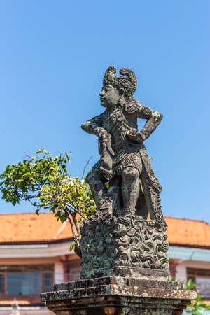 Klungkung, Bali, Indonesia - February 26, 2019: Closeup of Stone statue of defiant King covered in black mold against blue sky. Some green foliage and red roof.の写真素材