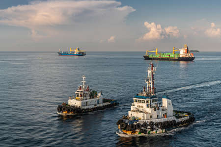 Makassar, Sulawesi, Indonesia - February 28, 2019: Black and white Numbay and Anoman tugboats , container and cargo ships outside de harbor on blue Flores Sea under early morning cloudscape.のeditorial素材
