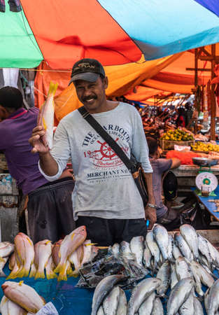 Makassar, Sulawesi, Indonesia - February 28, 2019: Terong Street Market. Smiling merchant shows one fish with a heap of more fishes lying in front of him. vegetable booth in back. Tarps of many colors.のeditorial素材