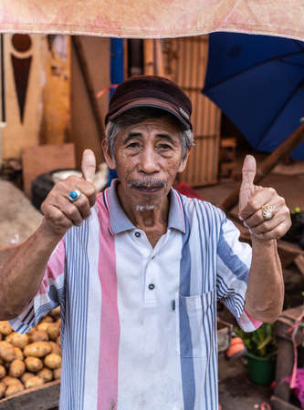 Makassar, Sulawesi, Indonesia - February 28, 2019: Terong Street Market. Portrait of male senior in white shirt and maroon cap flashing positive hand signals. Vegetable booth in back.のeditorial素材