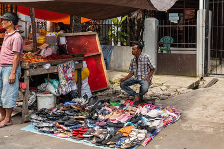 Makassar, Sulawesi, Indonesia - February 28, 2019: Terong Street Market. Ambulant male shoe vendor has spread a pile of shoes on a tarp on the streets dirt surface. Veggie booth nearby.のeditorial素材