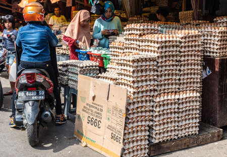 Makassar, Sulawesi, Indonesia - February 28, 2019: Terong Street Market. Closeup of booth with large stacks of cartons filled with brown eggs. Shoppers and guy on motorbike.のeditorial素材