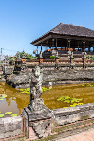 Klungkung, Bali, Indonesia - February 26, 2019: Brown stone Floating Pavilion at Royal Palace of Klungkung Kingdom with gray reed roof under blue sky. Mold covered monuments and yellow-brown lotus pond. Front is gray stone statue.のeditorial素材