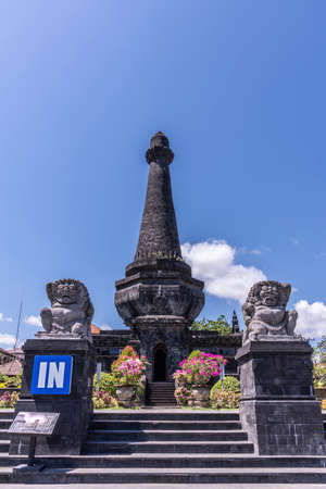 Klungkung, Bali, Indonesia - February 26, 2019: Black Oblisk Puputan Monument remembering a suicidal royal battle against invaders under blue sky. Phallus symbol and two lion statues as guards. Pink flowers set in green.のeditorial素材