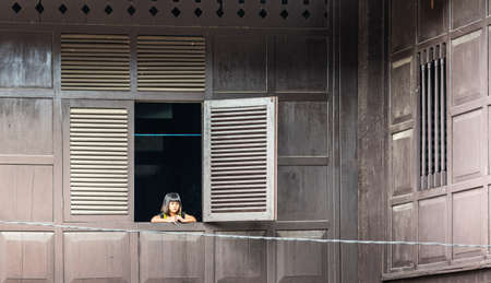 Makassar, Sulawesi, Indonesia - February 28, 2019: Little girl looks out of open window, set in dark...