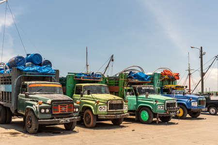 Makassar, Sulawesi, Indonesia - February 28, 2019: Paotere Old Port. Closeup of line of the front of four old Toyota trucks, backed up at the edge of the dock under blue sky. Greens, blues, darks, and reds.のeditorial素材