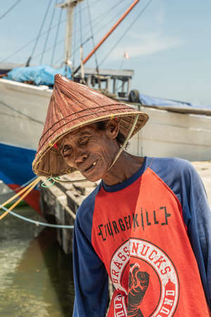 Makassar, Sulawesi, Indonesia - February 28, 2019: Paotere Old Port. Closeup of old man dock worker with cone straw hat and blue-red-white shirt.  Bow of boat on back and blue sky.のeditorial素材