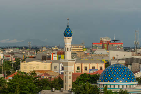 Makassar, Sulawesi, Indonesia - February 28, 2019: Masjid Babulssalam Pelabuhan mosque with dome and minaret in front of large city skyline.. Rain storn approaching cloudscape. Antennas on roofs.のeditorial素材