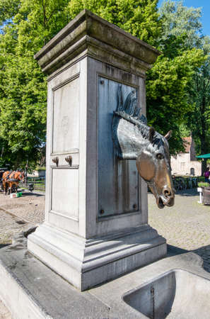 Bruges, Flanders, Belgium -  June 17, 2019: Gray stone water pump with horse head as faucet especially for horses of carriages near beguinage. Green foliage, brown horse in back.の写真素材