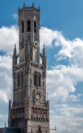 Bruges, Flanders, Belgium -  June 17, 2019: Closeup of Belfry tall gray brick clock tower, Halletoren, against blue sky with white clouds.の写真素材