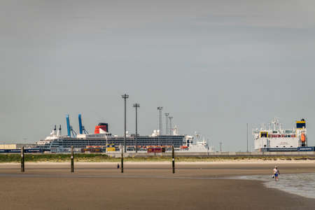 Zeebrugge, Flanders, Belgium -  June 18, 2019: Queen Mary 2 Cruise Ship in port of Zeebrugg as seen from beach in Knokke-Heist under gray-blue sky. Wet sand in front. Walkers.のeditorial素材